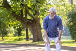 Tired senior gray-haired man is standing bent over in the park and resting after an active walk, run and workout